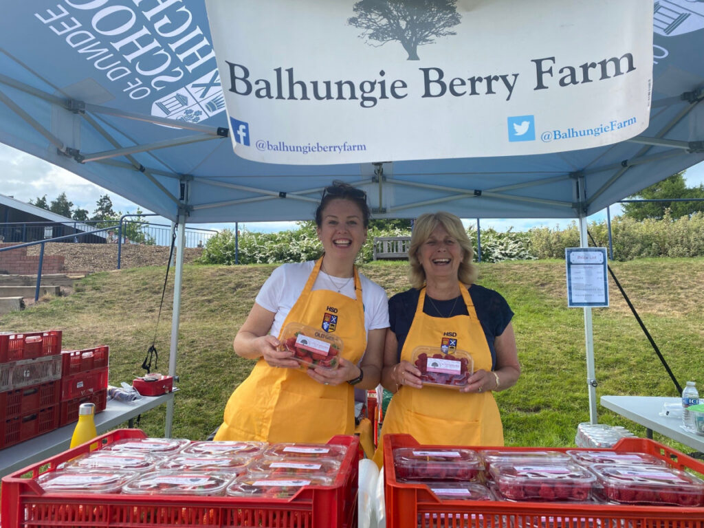 ALUMNI Old Girls. Associates of school at a strawberry stand stall surrounded by packed strawberries and holding them.