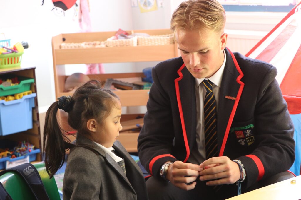Belonging - FB House badge presentations from F6 to L1s. Pupil getting presented a badge to a younger pupil in classroom.