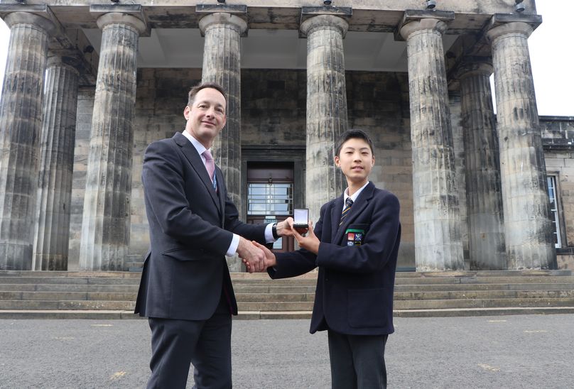 A Gold Standard Performance! A pupil being awarded a medal while shaking hands with a school staff member. Being presented to pupil outside front of school building.