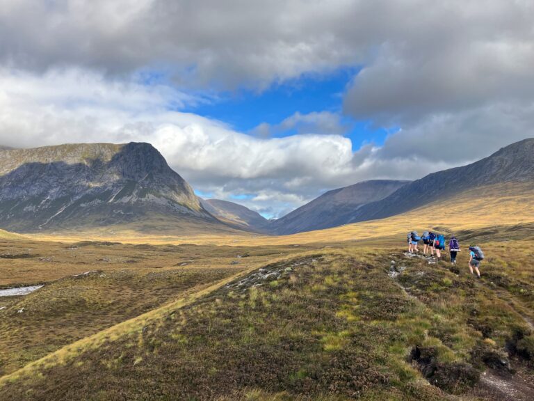 DofE 2. Pupils with rambling and camping gear on rambling through a meadow in valley with mountains surrounding them.