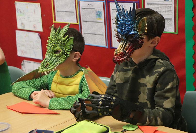 Dragon Day Is A Blazing Success!. Pupils in classroom sitting at desk, dressed up as dragons.