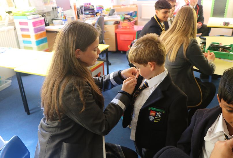 F6 Prefects Show L1 Pupils The Way. Pupils in classroom. Young pupil getting a badge pin put on his uniform by older pupil.