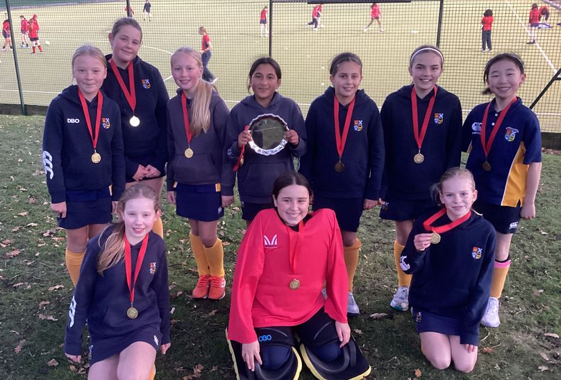 Hockey Silverware For L7 Girls. Pupils posing with awards of trophy and medals around their necks.All posing in line . Pupils in background playing hockey on courts.