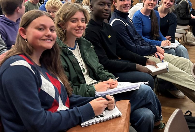 Success For Debaters In National Contest. Pupils sitting down in hall looking joyful in home clothes, with pen and notebooks.